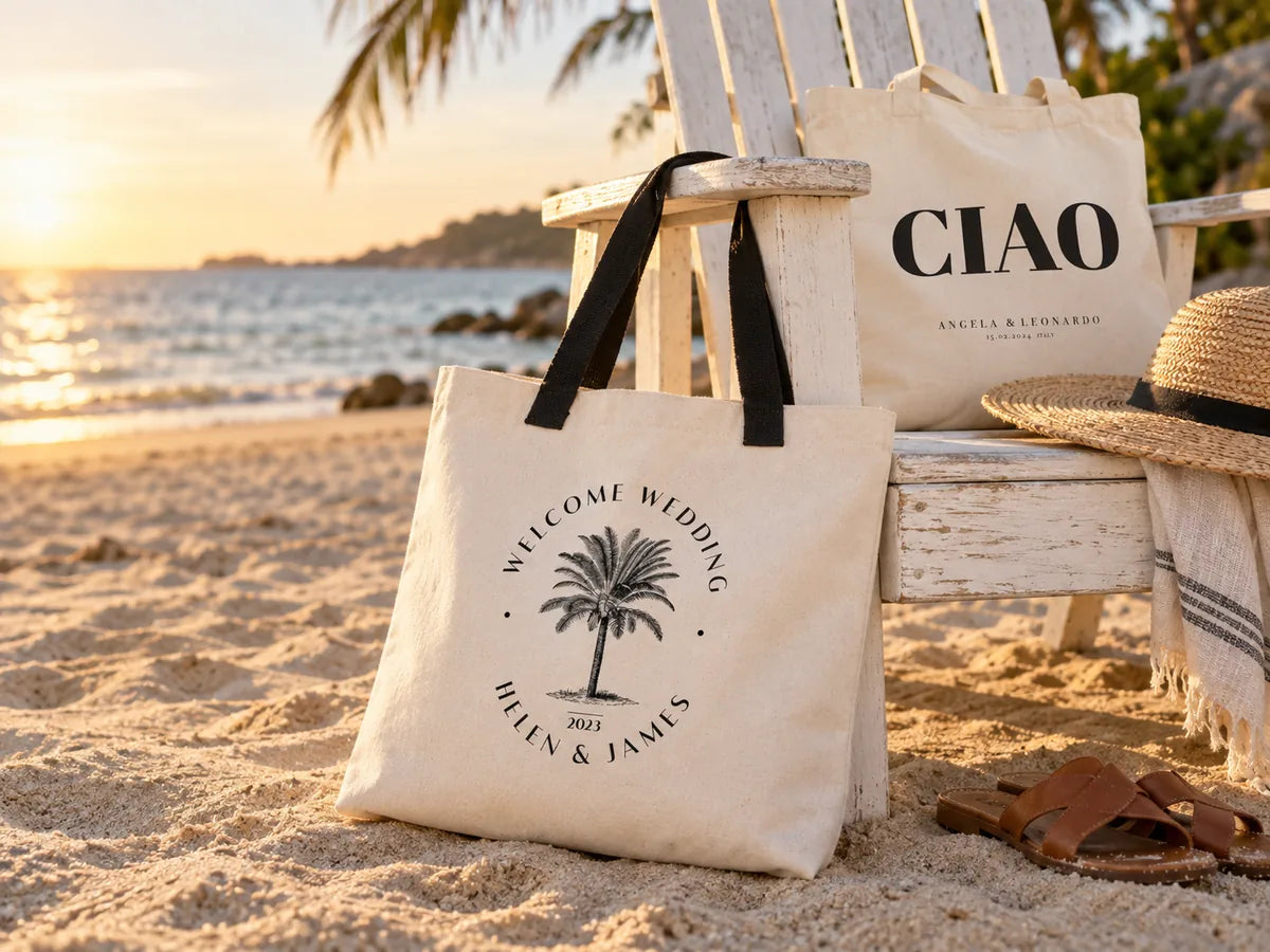 Spiaggia nell'ora dorata: grande tote bag in tela «Welcome Wedding Helen & James» con palma stampata e manici neri appoggiata a una sedia Adirondack bianca, dietro una tote «CIAO», un cappello di paglia e sandali di cuoio